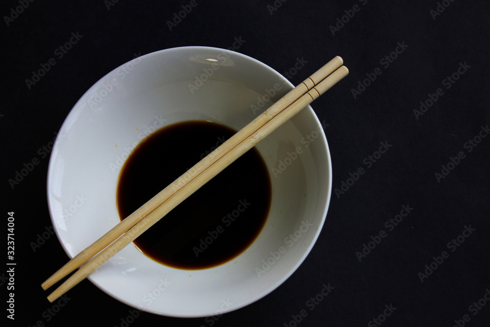 A white bowl of soy sauce and two chopsticks on a black background. A white bowl of soy sauce and two chopsticks on a black background.