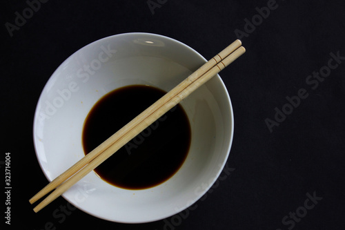 A white bowl of soy sauce and two chopsticks on a black background.