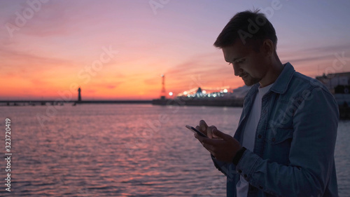 At sunset, a man uses a phone, the sea and the bright red sky in the background.