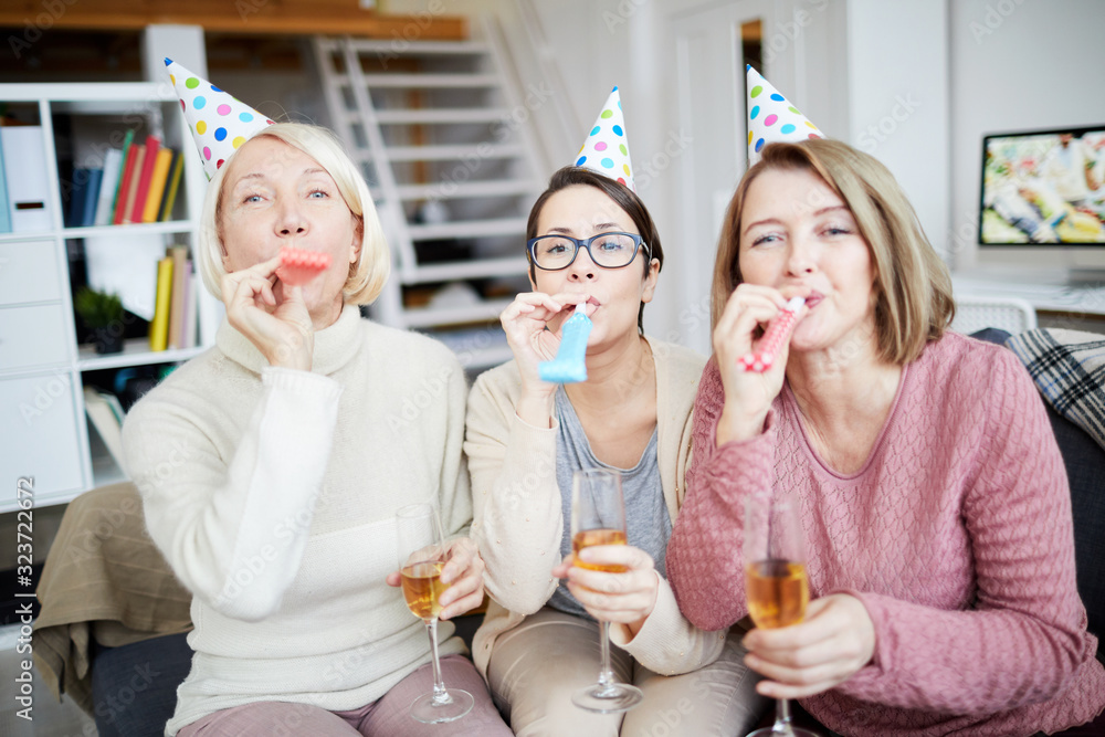 Portrait of three happy women blowing party horns and looking at camera while celebrating birthday at home - obrazy, fototapety, plakaty 