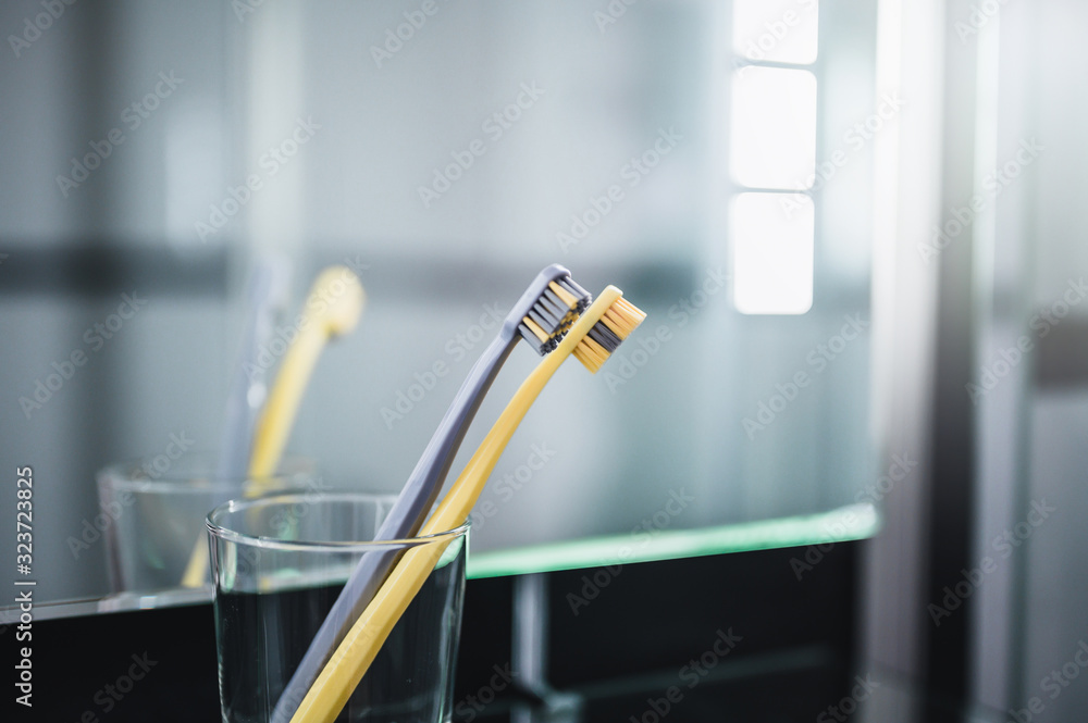 Two toothbrushes in glass cup in bathroom. Yellow and gray toothbrushes. Toothbrushes close up. Two toothbrushes in glass cup in bathroom. Yellow and gray toothbrushes. Toothbrushes close up.