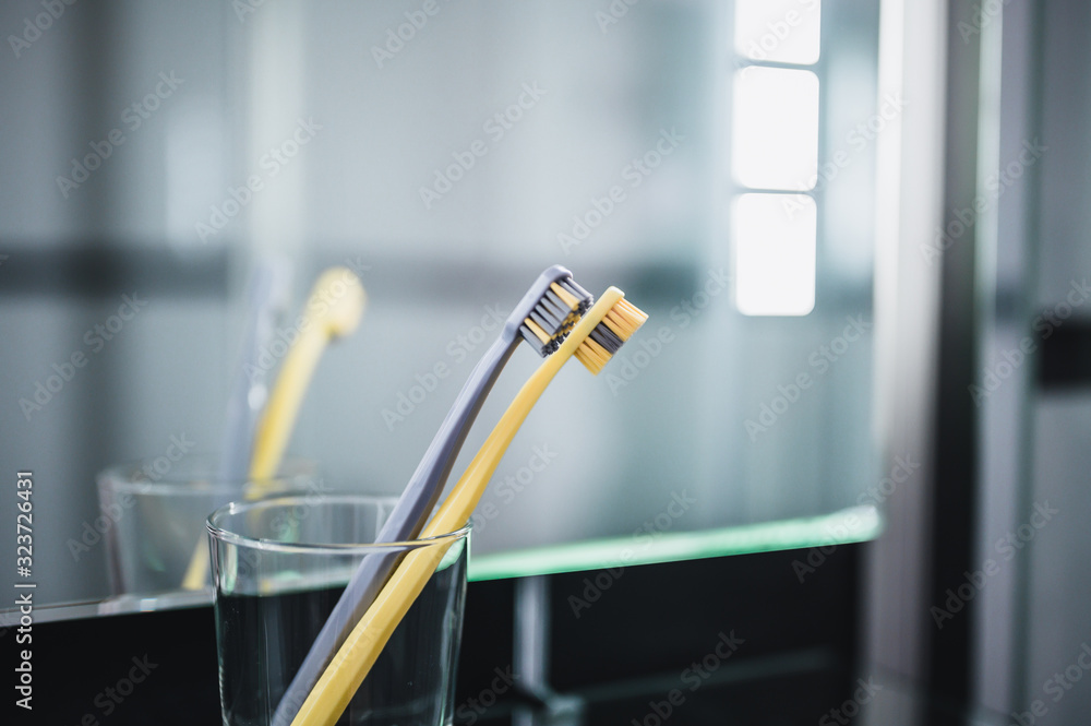 Two toothbrushes in glass cup in bathroom. Yellow and gray toothbrushes. Toothbrushes close up. Two toothbrushes in glass cup in bathroom. Yellow and gray toothbrushes. Toothbrushes close up.