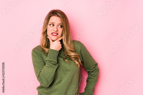 Young caucasian woman isolated on pink background touching back of head, thinking and making a choice.