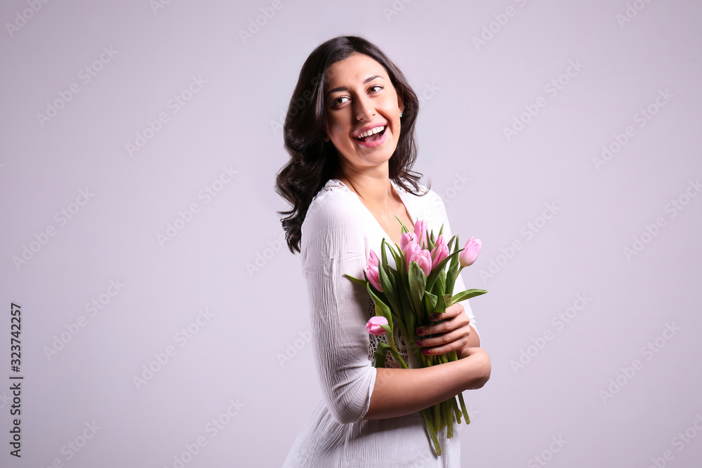 Studio portrait of gorgeous young brunette woman with long wavy hair wearing tight white wedding dress , holding bouquet of pink tulip flowers. Grey isolated background, copy space, close up. Studio portrait of gorgeous young brunette woman with long wavy hair wearing tight white wedding dress , holding bouquet of pink tulip flowers. Grey isolated background, copy space, close up.