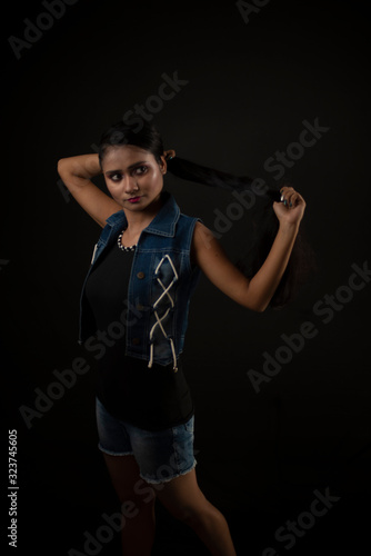 Fashion portrait of an young and attractive Indian Bengali brunette girl with tee shirt and blue western jacket tying hair in front of a black studio background. Indian fashion portrait and lifestyle.