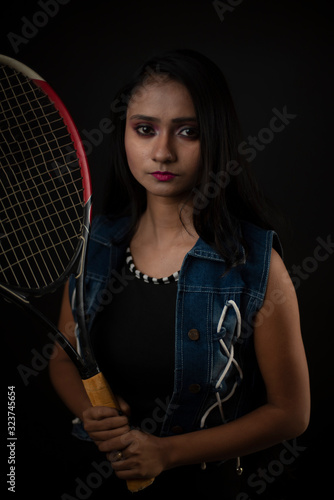 Fashion portrait of an young and attractive Indian Bengali brunette girl with tee shirt, blue western jacket and racket in front of a black studio background. Indian fashion portrait and lifestyle. © abir
