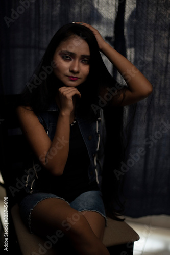 Fashion portrait of an young and attractive Indian Bengali brunette girl with tee shirt and blue western jacket sitting in front of a black studio background. Indian fashion portrait and lifestyle.
