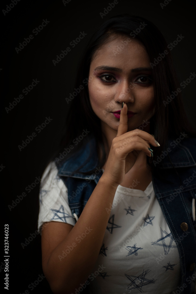 Obraz premium Fashion portrait of an young Indian Bengali brunette girl with tee shirt and blue western jacket putting finger on lips in front of a black studio background. Indian fashion portrait and lifestyle.