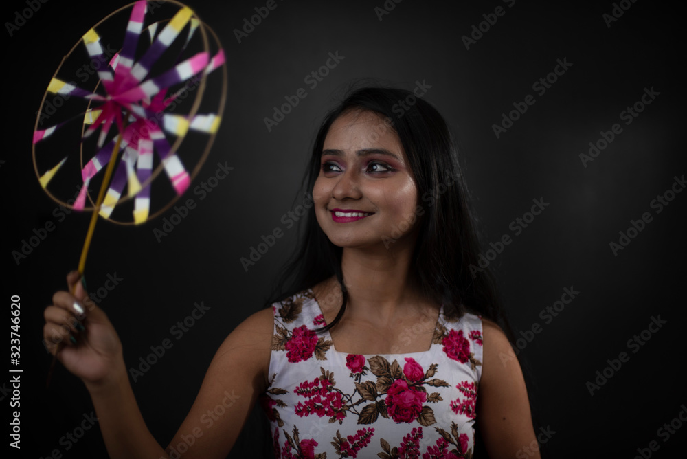 Fototapeta premium Fashion portrait of an young and attractive Indian Bengali brunette girl with floral western dress with pinwheel in front of a black studio background. Indian fashion portrait and lifestyle.