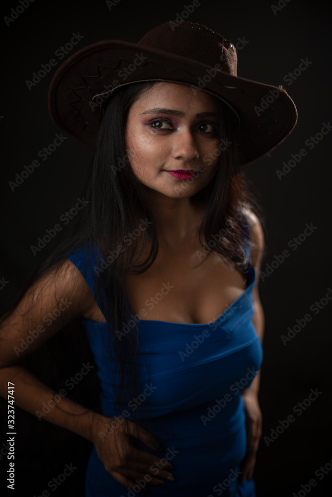 Fashion portrait of young and attractive Indian Bengali brunette girl with blue western dress with cowboy hat under spotlight in front of black studio background.Indian fashion portrait and lifestyle. Fashion portrait of young and attractive Indian Bengali brunette girl with blue western dress with cowboy hat under spotlight in front of black studio background.Indian fashion portrait and lifestyle.