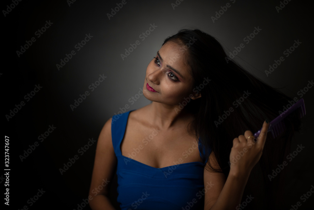 Fototapeta premium Fashion portrait of young Indian Bengali brunette girl with blue western dress combing her hair looking at mirror in front of black studio background. Indian fashion portrait and lifestyle.