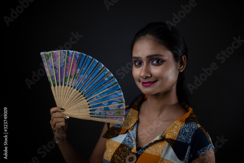 Fashion portrait of an young and attractive Indian Bengali brunette girl with checkered western dress holding Chinese fan in front of a black studio background. Indian fashion portrait and lifestyle.