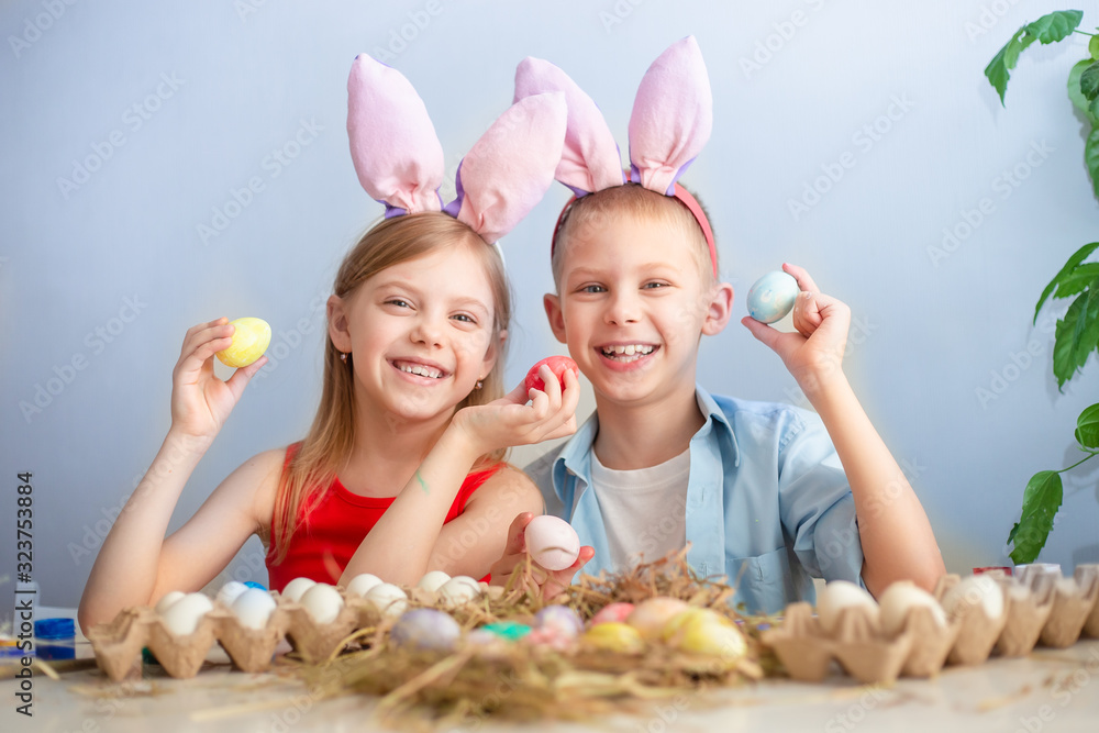 cute smiling children, fair-haired brother and sister of 7-9 years old, wear bunny ears on their heads and paint Easter eggs on a blue background cute smiling children, fair-haired brother and sister of 7-9 years old, wear bunny ears on their heads and paint Easter eggs on a blue background