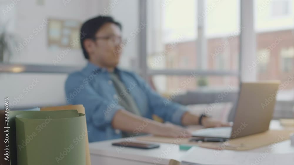 Selective focus shot of rolled paper in office and Asian architect using laptop and getting upset about project while working at desk in the background Selective focus shot of rolled paper in office and Asian architect using laptop and getting upset about project while working at desk in the background
