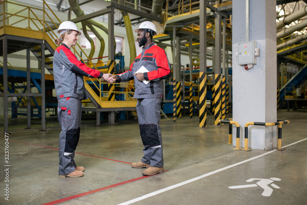 Fototapeta premium Horizontal shot of two factory engineers wearing uniform greeting each other with handshake