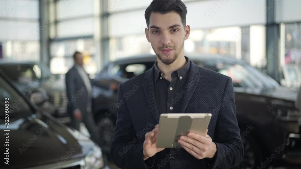 Young handsome brown-eyed Caucasian man standing in car dealership with tablet, looking at camera and smiling. Confident brunette dealer posing at workplace in showroom. Job, occupation, lifestyle. Young handsome brown-eyed Caucasian man standing in car dealership with tablet, looking at camera and smiling. Confident brunette dealer posing at workplace in showroom. Job, occupation, lifestyle.
