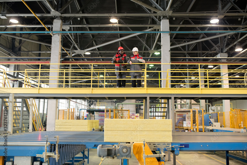 Horizontal long shot of two construction supplies factory workers wearing uniform standing together talking about something