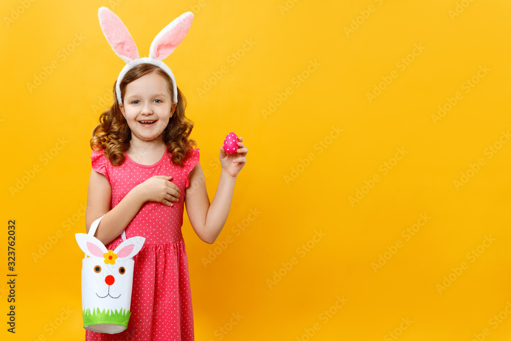 A beautiful cute little girl in the ears of a bunny holds a bag for sweets and an avshalny egg. Portrait of a child on a yellow background. Copy space