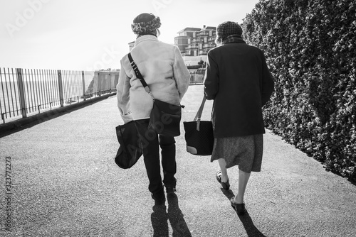 Two senior ladies walk along a cliff top promenade into the sunshine, in winter, chatting. They are seen from behind. There is some lens flare. The image is in monochrome photo