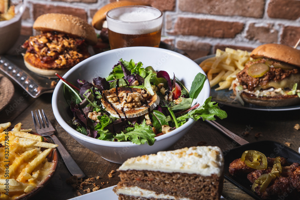 Variety of dishes, salad with goat cheese homemade hamburgers and french fries, chicken wings with jalapeños drink and cake on the wooden table. Isolated image. Variety of dishes, salad with goat cheese homemade hamburgers and french fries, chicken wings with jalapeños drink and cake on the wooden table. Isolated image.