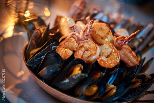 A full luxury dinner ceramic bowl of prepared grilled shrimps and mussels including tiny squids served on a white stone table next to golden candle in a soft light of a restaurant, close up