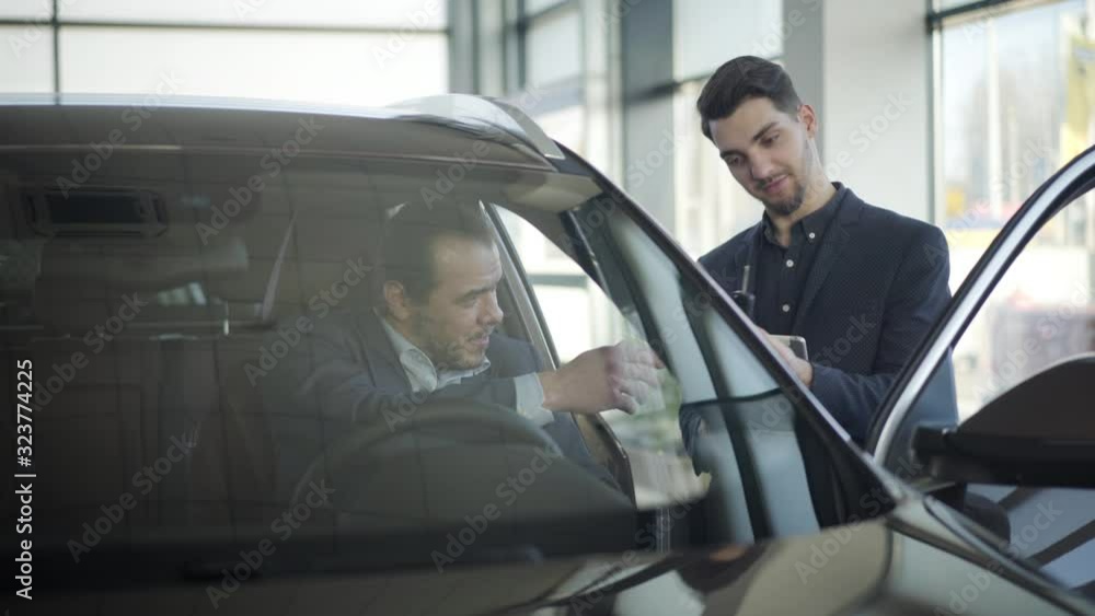 Smiling male Caucasian trader giving car keys to adult businessman sitting on driver's seat. Successful client buying automobile in dealership. Business, lifestyle, wealth, success. Smiling male Caucasian trader giving car keys to adult businessman sitting on driver's seat. Successful client buying automobile in dealership. Business, lifestyle, wealth, success.