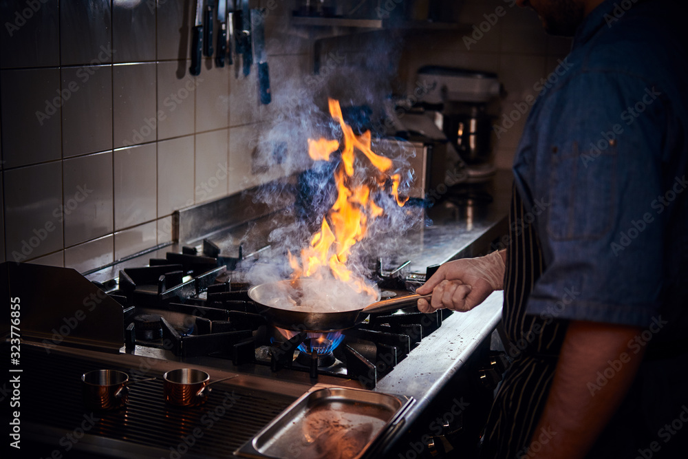 Professional chef wearing gloves and apron cooking stir-fry flambe on a pan with open fire in a dark restaurant kitchen Professional chef wearing gloves and apron cooking stir-fry flambe on a pan with open fire in a dark restaurant kitchen