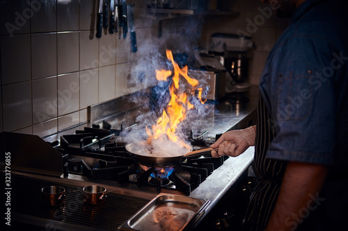 Professional chef wearing gloves and apron cooking stir-fry flambe on a pan with open fire in a dark restaurant kitchen