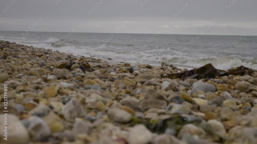 A zooming, extreme-closeup shot of different sizes of stones and pebbles on a rough and rocky shore amid overcast skies with waves crashing in the background. A zooming, extreme-closeup shot of different sizes of stones and pebbles on a rough and rocky shore amid overcast skies with waves crashing in the background.