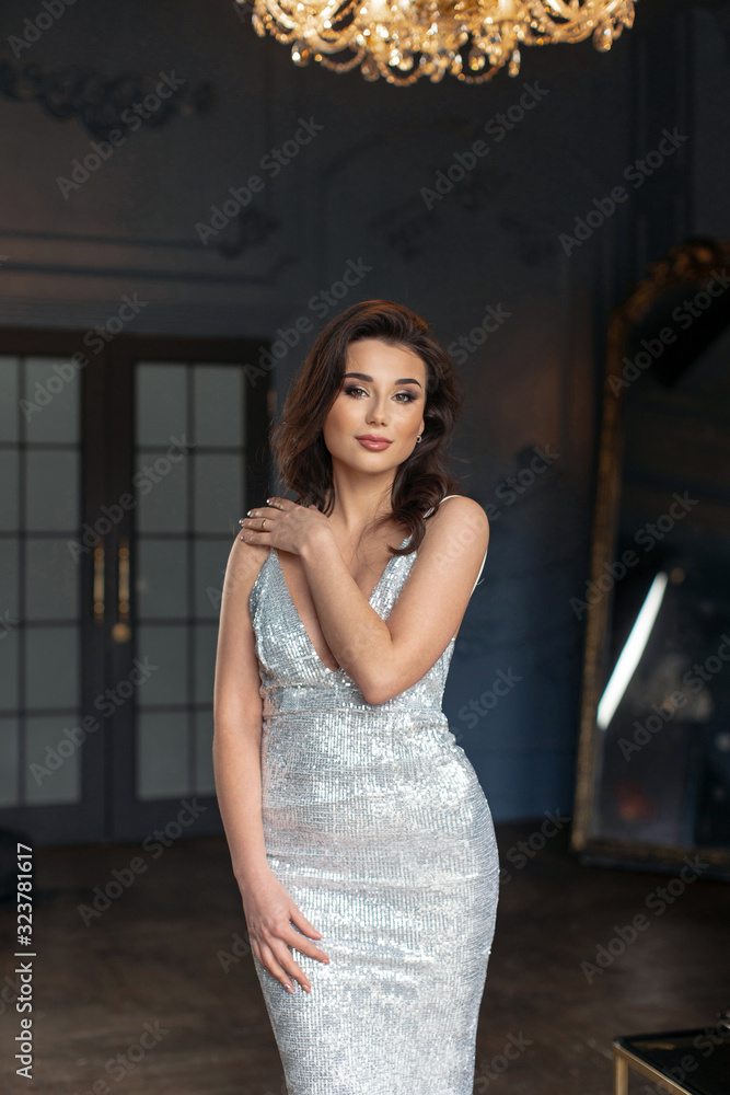 Sexy brunette with makeup in evening silver dress posing in deep blue vintage interior