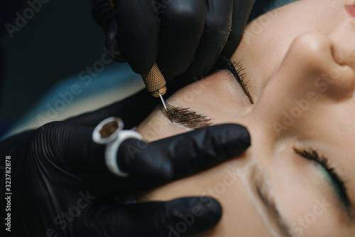 Hands in black gloves hold the manipulator with the microblading needle and draw her hair on the girl’s eyebrows close-up. photo