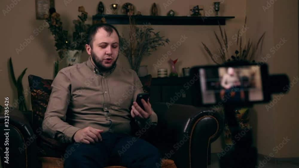The influential Bearded Young guy, a blogger, of European appearance, sits in a chair, records a video vlog and answers followers questions. Social Media Blogging Concept The influential Bearded Young guy, a blogger, of European appearance, sits in a chair, records a video vlog and answers followers questions. Social Media Blogging Concept