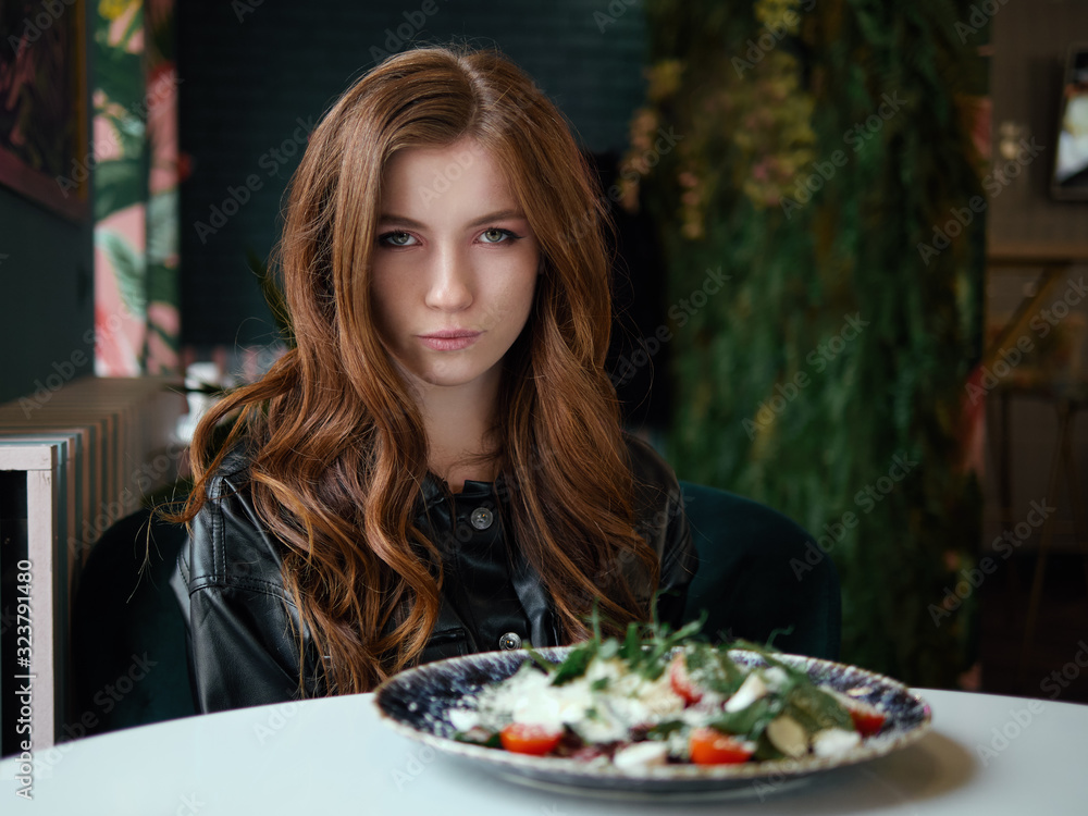 Beautiful not impressed redhead woman in black leather shirt sitting in trendy cozy city cafe near big window with delicious salad on table