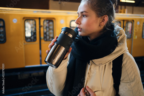 Side view of casual tourist girl on platform drinking warm beverage while waiting train in metro station