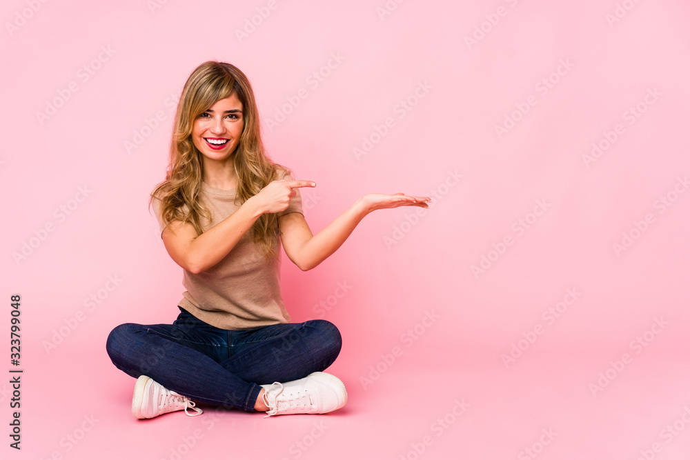 Young blonde caucasian woman sitting on a pink studio excited holding a copy space on palm. Young blonde caucasian woman sitting on a pink studio excited holding a copy space on palm.