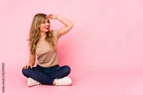 Young blonde caucasian woman sitting on a pink studio looking far away keeping hand on forehead.