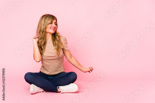 Young blonde caucasian woman sitting on a pink studio dancing and having fun.