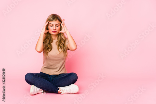 Young blonde caucasian woman sitting on a pink studio touching temples and having headache.