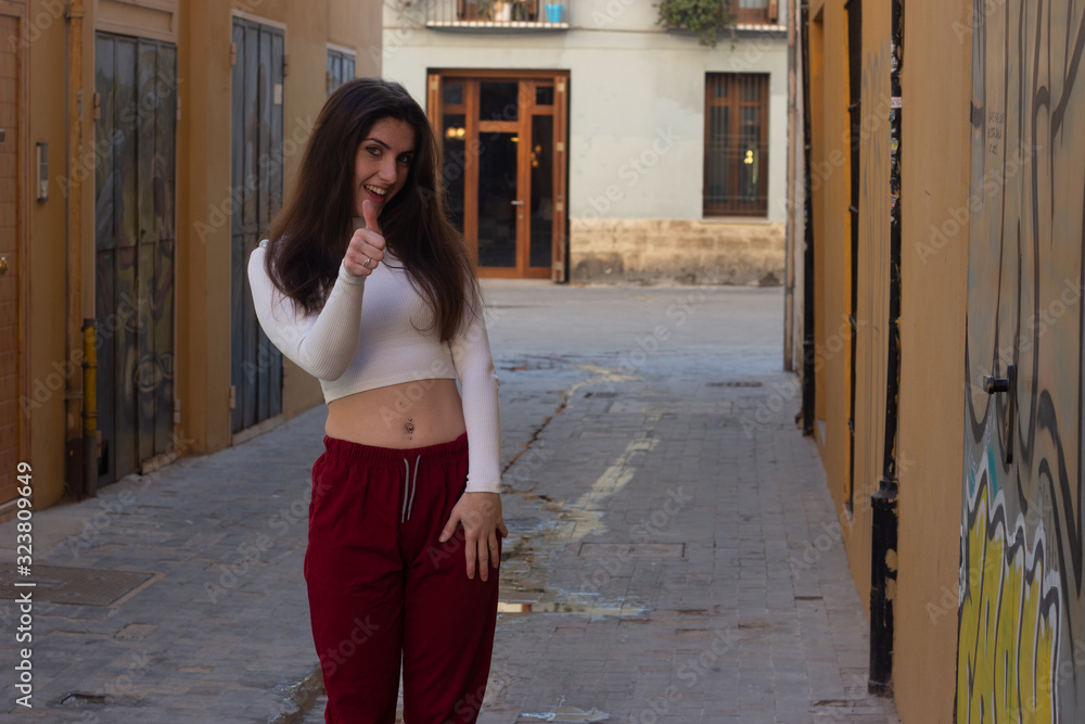 attractive young woman showing thumb up gesture, in the city, white top and red pants. Place for your text in copy space.