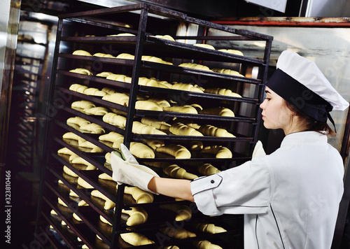woman Baker delivers a cart with pans of raw croissants to a large oven against the background of a bakery or bread factory.