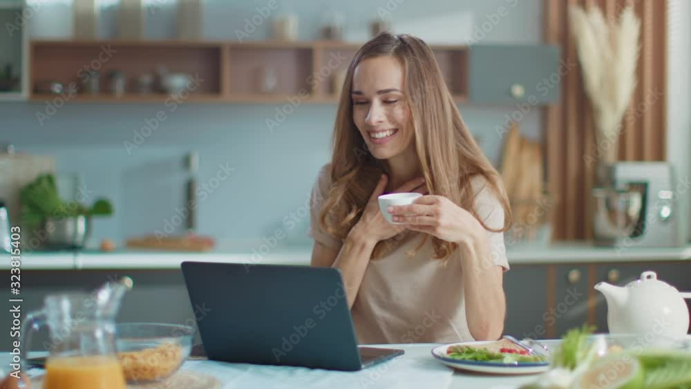 Woman chatting on webcam with friend on laptop at kitchen. Lady drinking coffee Woman chatting on webcam with friend on laptop at kitchen. Lady drinking coffee