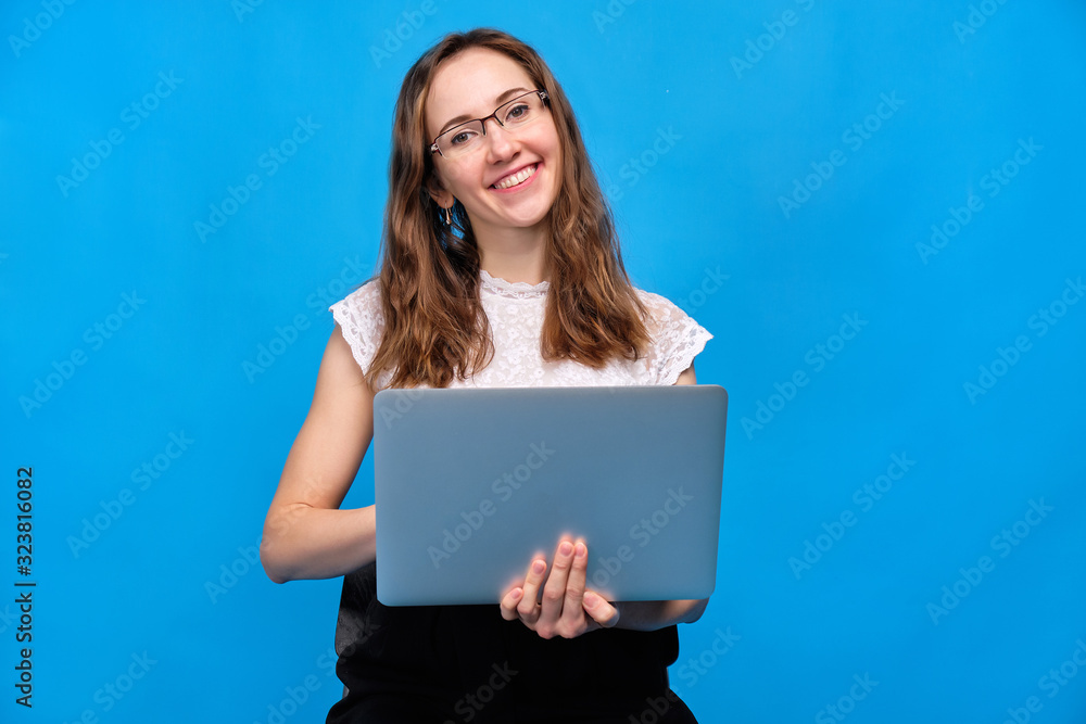 A brunette girl in casual clothes is holding a laptop on a blue background with glasses holding a laptop and looking at the camera. A brunette girl in casual clothes is holding a laptop on a blue background with glasses holding a laptop and looking at the camera.