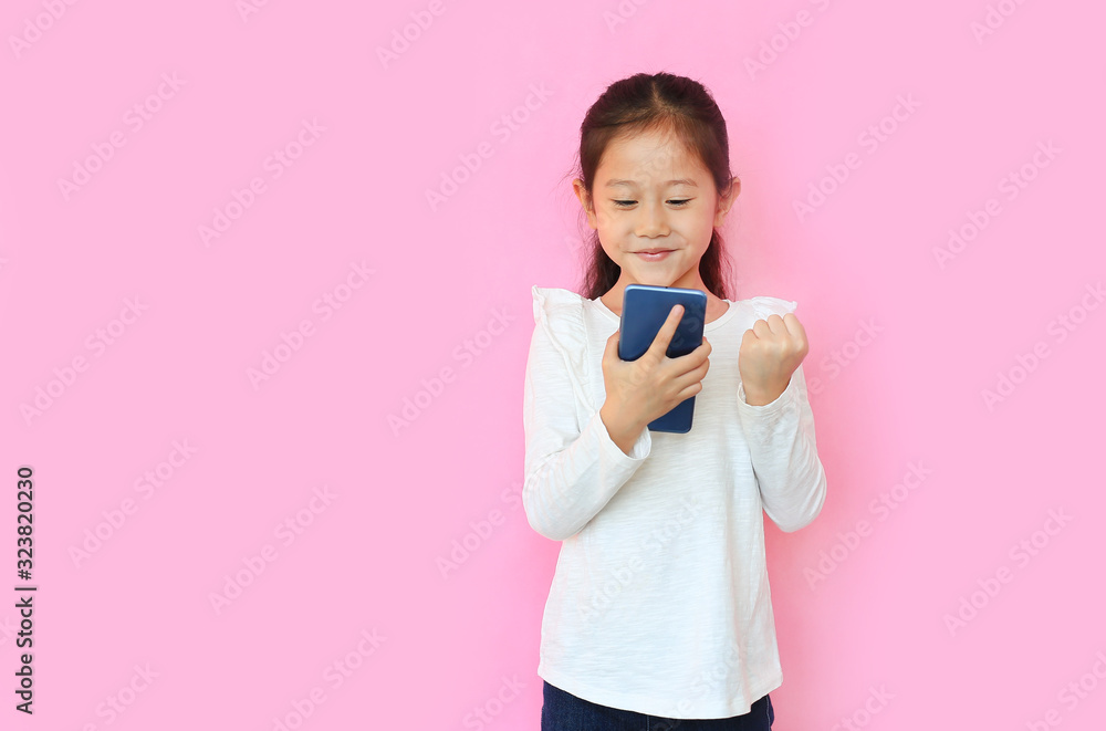 Surprised happy asian little child girl watching her smartphone and expression hand fight sign over pink background with copy space. Kid smiling and enjoy while playing cellphone. Surprised happy asian little child girl watching her smartphone and expression hand fight sign over pink background with copy space. Kid smiling and enjoy while playing cellphone.
