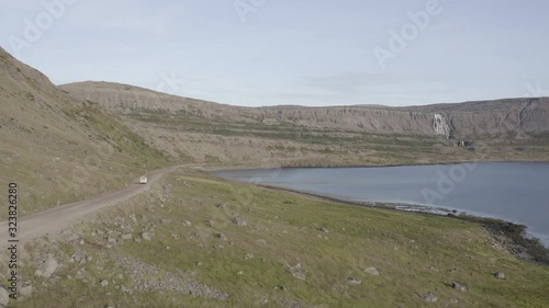 Beautiful aerial shot of a white car driving by the sea on a dusty road in north-west Iceland, the drone is following from a wide shot, with the Dynjandi waterfall in the background. photo