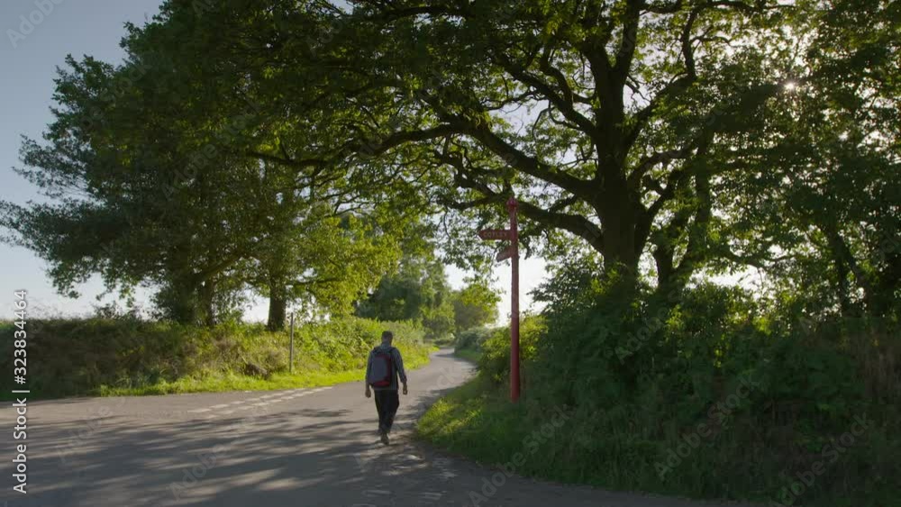 A daylight wide shot of a traveller carrying a backpack and walking down a curved paved road lined by trees and lush foliage. A daylight wide shot of a traveller carrying a backpack and walking down a curved paved road lined by trees and lush foliage.