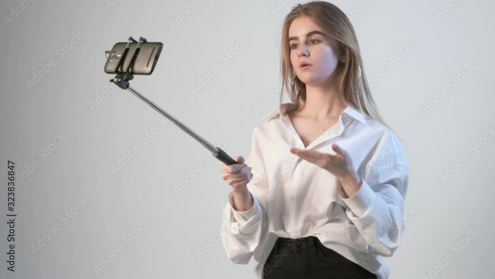 young female records video with smartphone attached to selfie stick. blond girl filming tutorial vlog about eyebrows cosmetics and makeup, using cellphone isolated on white background. Beauty blogger young female records video with smartphone attached to selfie stick. blond girl filming tutorial vlog about eyebrows cosmetics and makeup, using cellphone isolated on white background. Beauty blogger