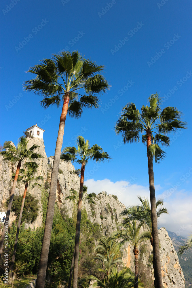white bell tower of castell de guadalest with palms on bright blue sky background, Spain