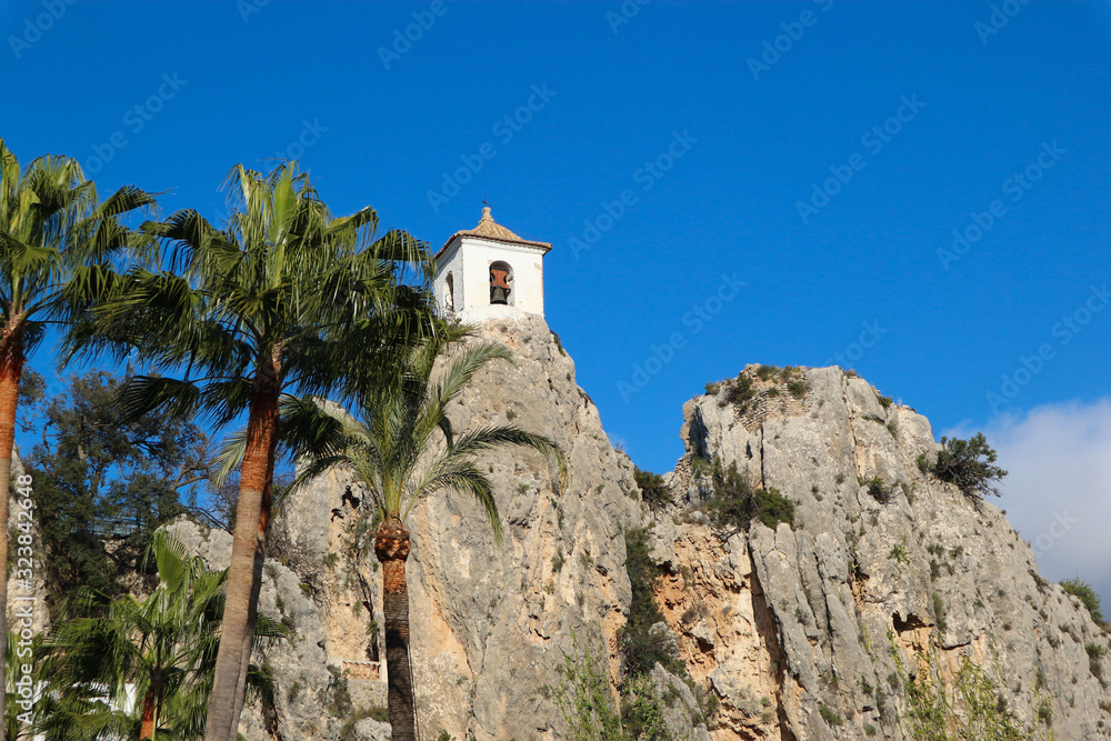 white bell tower of castell de guadalest with palms on bright blue sky background, Spain