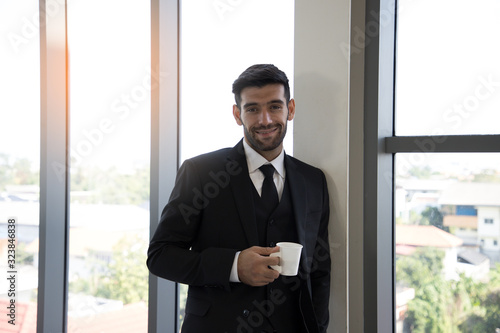 Portrait of a Handsome young businessman with a friendly and smiling broadly at the camera with self-assured expression while holding coffee cup and playing smartphone in the office. Relax time.