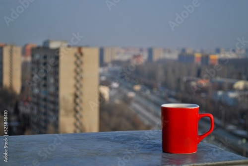 A red mug with black coffee stands on the parapet of the roof against the background of a winter panorama of the city in the morning sun.